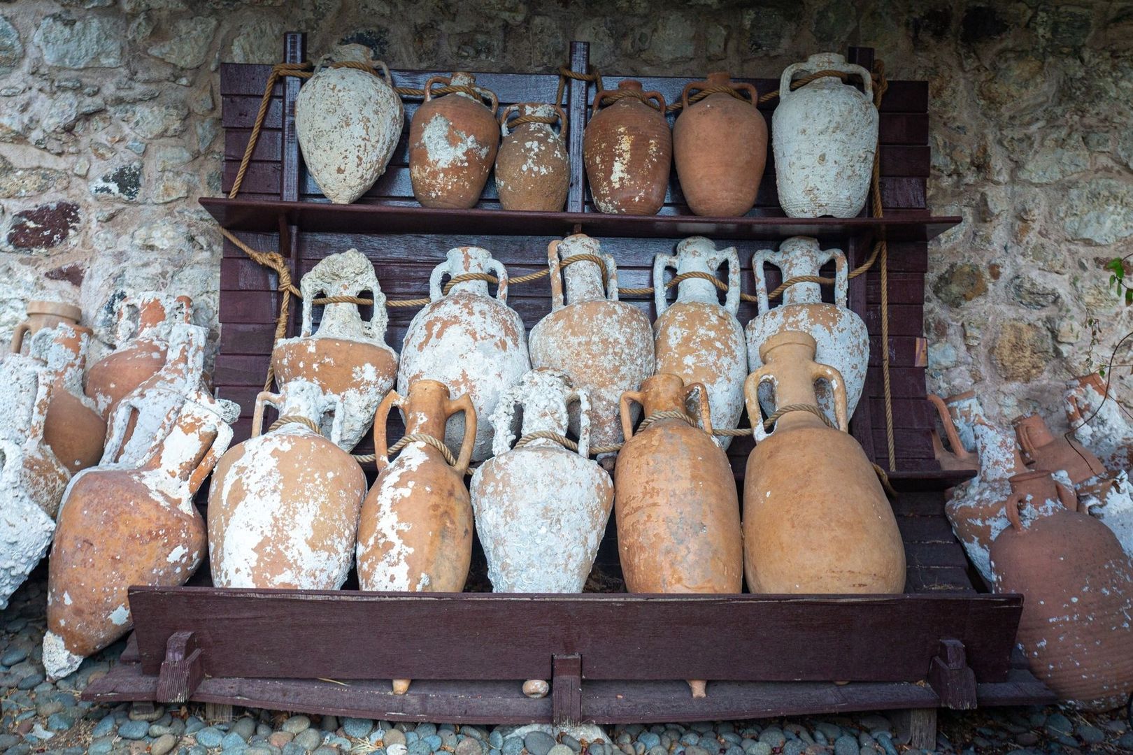 Weathered terracotta amphorae displayed on wooden racks against a stone wall